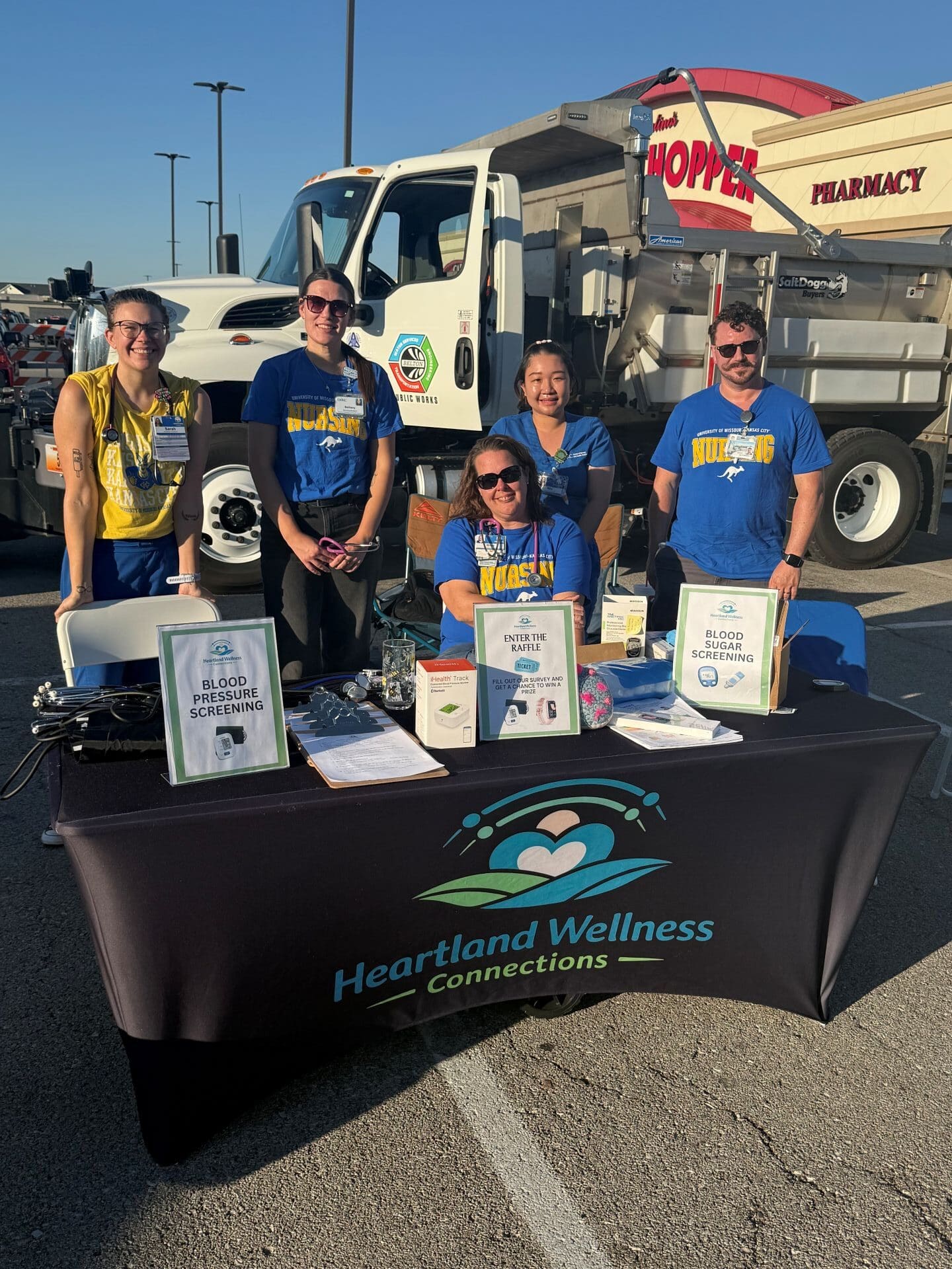 A group of people standing behind a table with the Heartland Wellness Connections logo printed on the tablecloth. A large truck is behind them.